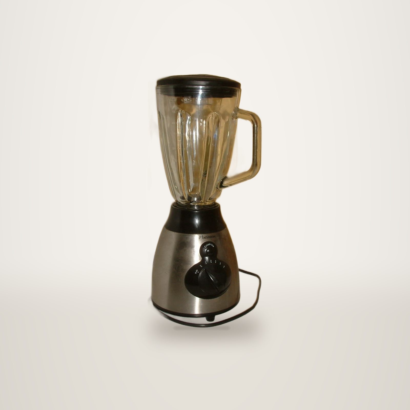 Un blender électrique avec un socle en inox brossé, un bouton de réglage noir et un grand bol en verre à anse, posé devant un mur clair.