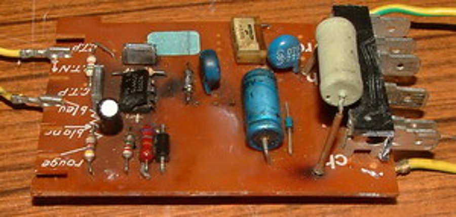 Circuit imprimé marron avec condensateurs bleus, résistance blanche et fils jaunes posé sur une surface en bois.