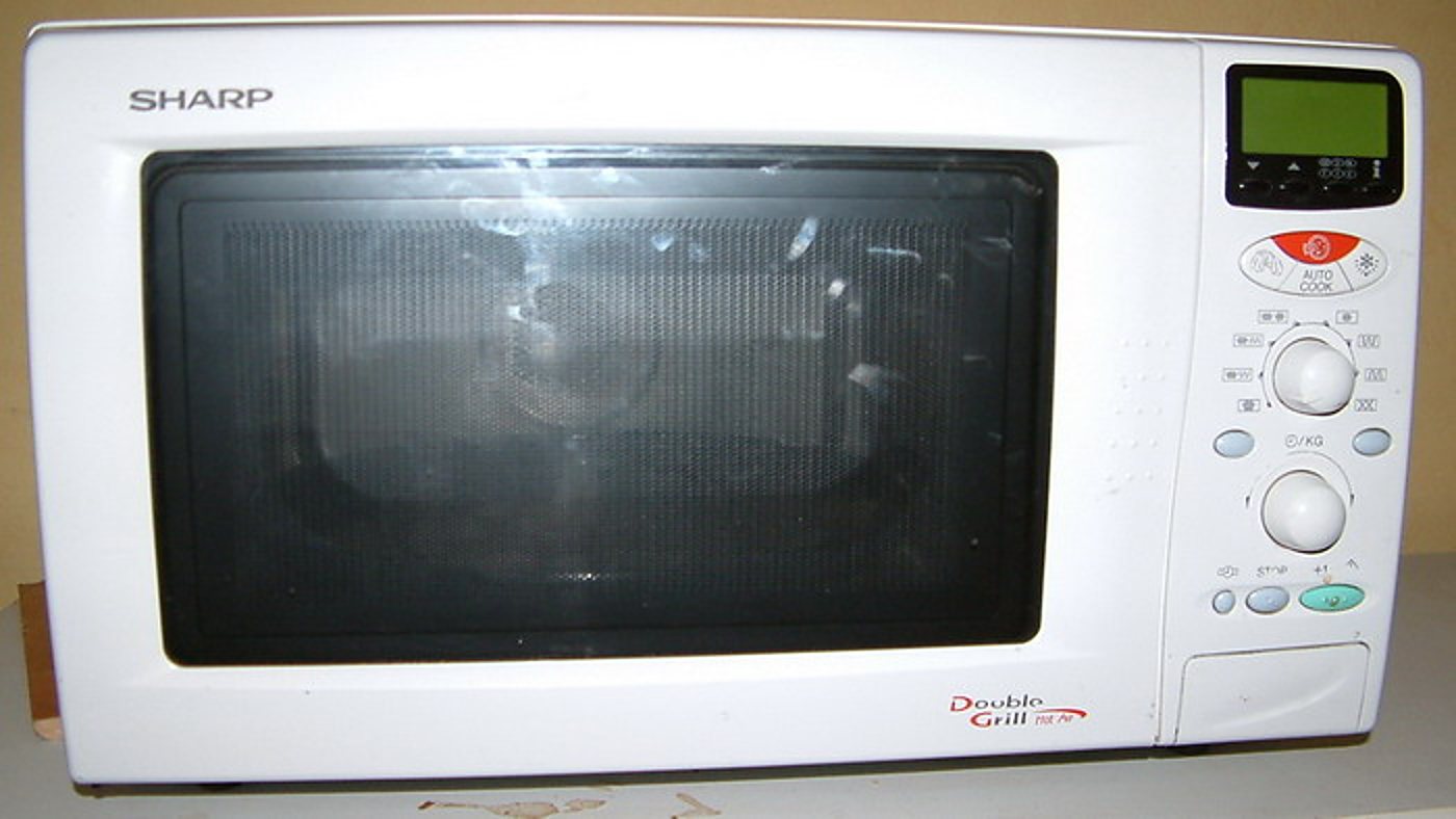Un four à micro-ondes Sharp blanc avec porte vitrée sombre, écran numérique vert et deux boutons de réglage, posé devant un mur beige.