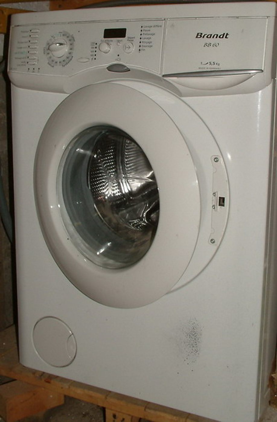 Un lave-linge blanc de marque Brandt à hublot rond, vu de trois quarts, posé sur une structure en bois dans un local.