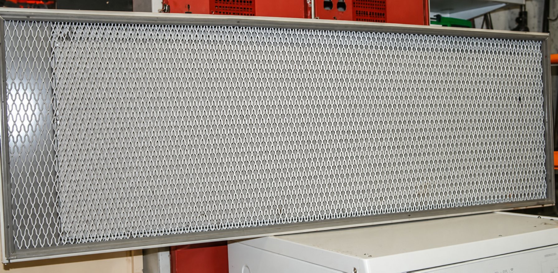 Un grand panneau rectangulaire à cadre beige et grillage métallique en losanges est posé sur un appareil blanc dans un atelier.