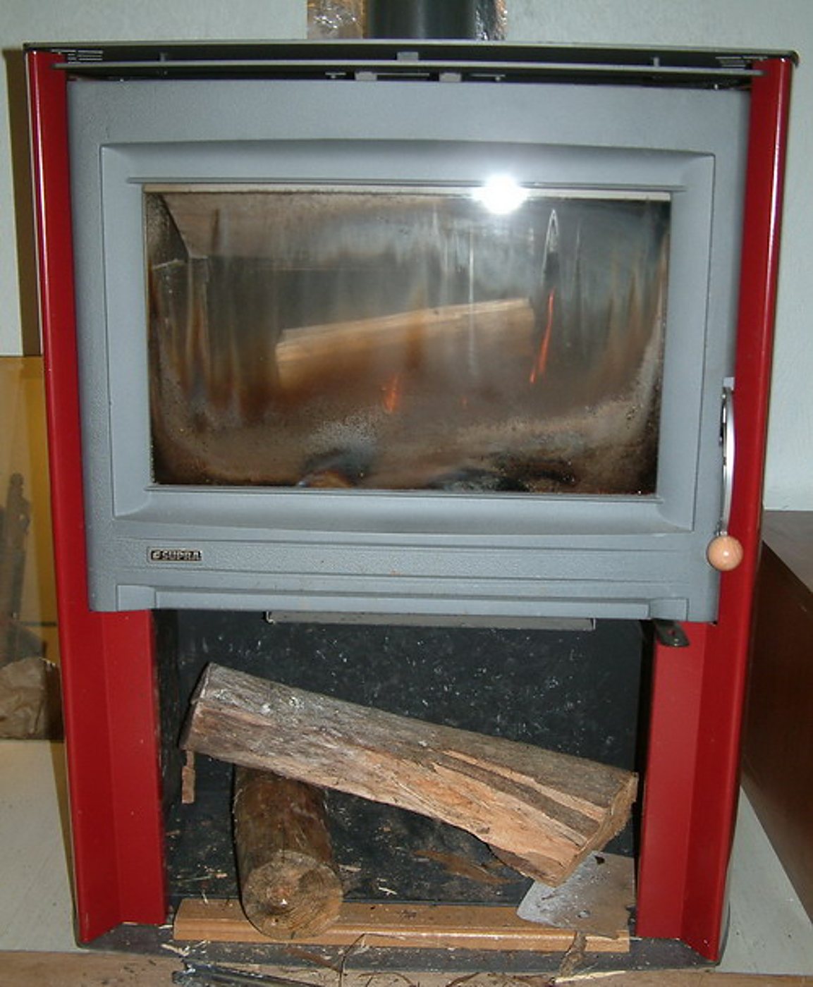 Un poêle à bois rouge et gris avec une grande vitre noircie, et deux bûches rangées dans le compartiment sous le foyer.