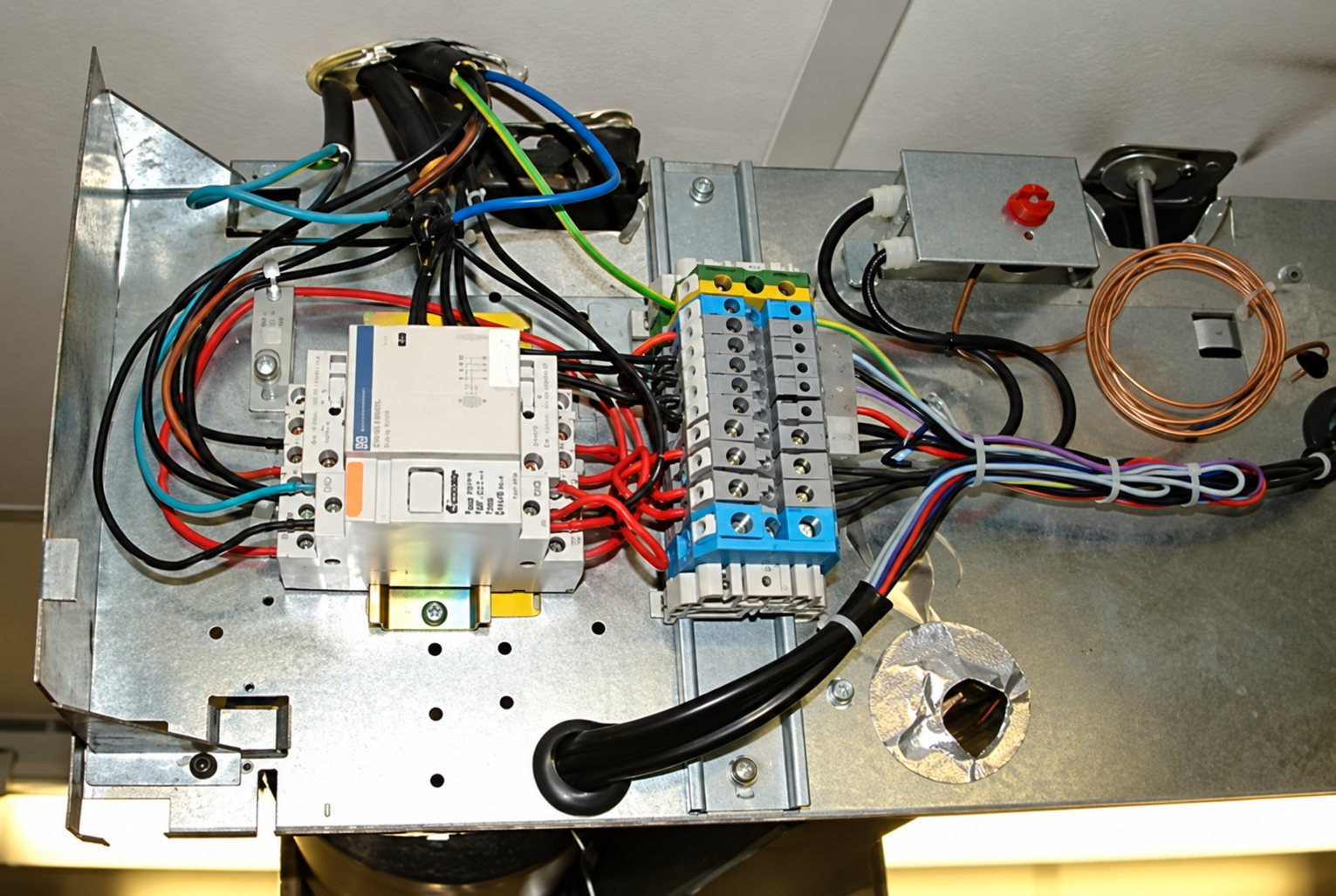 Vue de dessus d'un coffret électrique ouvert montrant des fils colorés, un disjoncteur modulaire et un bornier fixés sur une plaque métallique.
